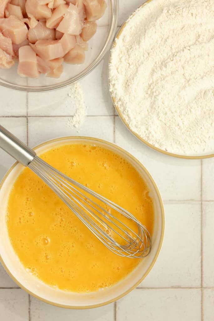 Chunks of chicken in a bowl with a bowl of flour and an egg mixture next to it.