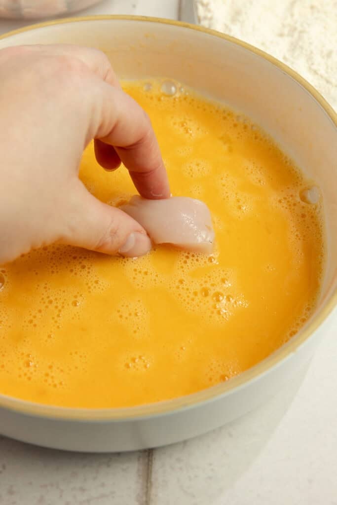 A hand dipping chicken into a bowl of whisked egg.