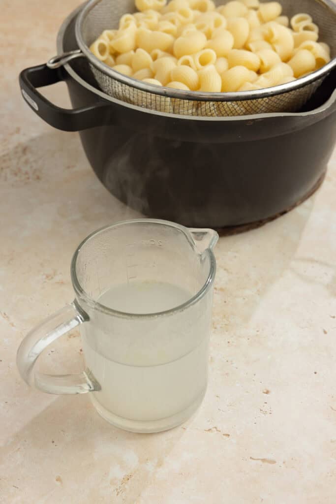 A pot with a strainer inside it full of pasta next to a glass pouring dish with pasta water.